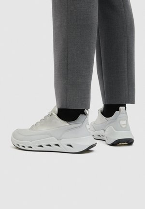 Person iført hvide læder sneakers med ventilerede såler og grå bukser, stående på en ensfarvet hvid baggrund.