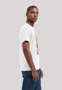Weißes T-Shirt mit kurzen Ärmeln, das ein farbenfrohes Grafikdesign auf der Vorderseite zeigt. Das Modell trägt dunkelblaue Jeans und steht vor einem neutralen Hintergrund.