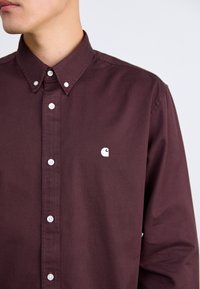 Camisa botones burdeos con cuello clásico, con botones blancos y un pequeño logo blanco en el lado izquierdo del pecho. Textura de algodón suave.