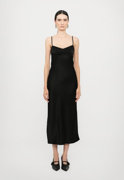 Vestido de satén negro con tirantes finos, que presenta un cuerpo ajustado y una falda fluida. Combinado con zapatos negros de punta con lazos.