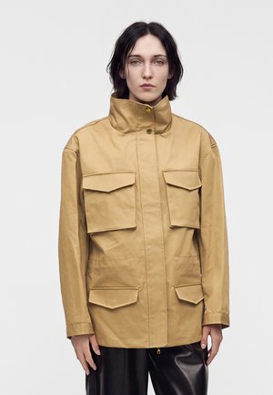 Personne aux cheveux courts et foncés porte une veste beige à col haut avec quatre poches avant à rabat, associée à un pantalon noir, debout devant un fond uni.