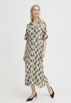 Nő zöld-fehér mintás, gombos, midi hosszúságú ruhában, rövid ujjúval és fekete pántos magas sarkú cipőben, fehér háttér előtt állva.
