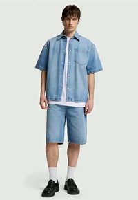 Camisa de azul claro em denim de mangas curtas com um bolso frontal, usada sobre uma t-shirt branca e combinada com calções de denim a condizer e sapatos pretos.