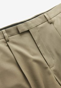 Pantaloni beige su misura con una texture liscia, caratterizzati da pieghe frontali, vita piatta e dettagli di cucitura sottili.