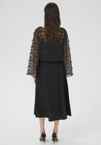 Sieviete stāv ar muguru pret skatītāju, tērpusies melnā caurspīdīgā mežģīņu blūzē ar garām piedurknēm pāri melnai kleitai un melnām augstpapēžu kurpēm uz baltas fona.