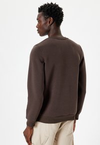 Sudadera de algodón marrón con un cuello redondo y puños acanalados, mostrada desde atrás. La textura parece suave y tiene un corte holgado.