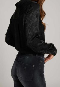 Kvinde iført sort tekstureret langærmet skjorte, stoppet ned i tætsiddende mørk denim jeans med baglommer og et lille logo patch.