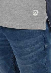 Szürke pamut ing oldalsó hasítékokkal, fehér hímzett logóval. Sötétkék farmernadrággal párosítva, amelyen világos kopások láthatók.