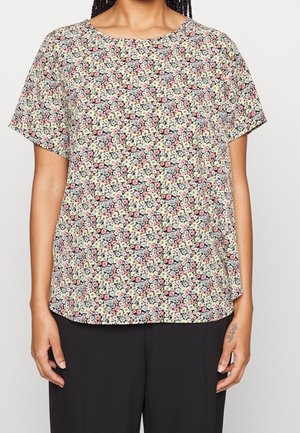 Kobieta ubrana w krótki rękaw bluzki w kwiaty z małymi różowymi, białymi i szarymi kwiatami, zestawioną z czarnymi spodniami.