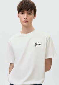 Valkoinen puuvillainen t-paita lyhyillä hihoilla, jossa on musta brodeerattu "Fender"-logo vasemmassa rinnassa. Yksinkertainen pyöreä kaula-aukko.