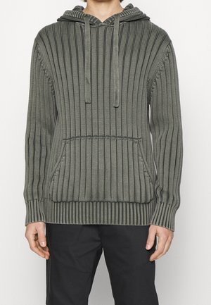 Personne portant un sweat à capuche en maille grise côtelée avec poche avant et cordons, associé à un pantalon noir, debout devant un fond blanc uni.
