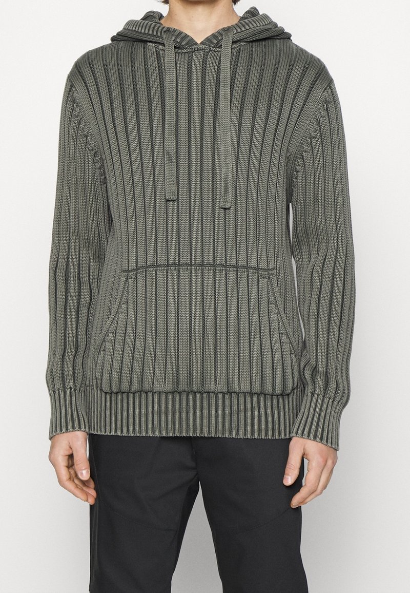 Personne portant un sweat à capuche en maille grise côtelée avec poche avant et cordons, associé à un pantalon noir, debout devant un fond blanc uni.