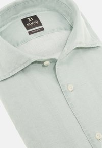 Camicetta di lino di colore verde chiaro con una texture morbida, frontale con bottoni e colletto classico. L'etichetta mostra il nome del marchio e informazioni sulla vestibilità.