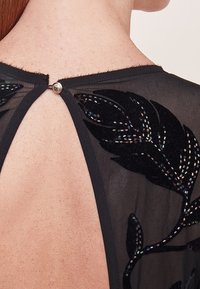 Černá transparentní tkanina s květinovým vzorem, zdobená korálky. Zapínání na zádech pomocí ozdobného knoflíku na krku.