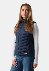 Junge Frau mit langen Haaren, die eine gezippte marineblaue gesteppte Weste über einem weißen Langarmshirt und blauen Jeans trägt, vor weißem Hintergrund stehend.