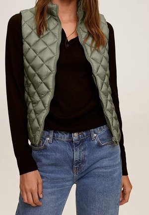 Kvinde iført en lysegrøn, quiltet vest over en sort langærmet skjorte og blå denimjeans, stående med den ene hånd afslappet ved siden.