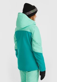 Teal en lichtgroene skijas met een capuchon, elastische manchetten en contrasterende details. Draag het met zwarte handschoenen en een beanie.