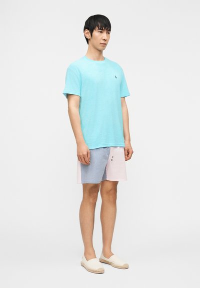 Νεαρός άνδρας που στέκεται με τα χέρια στα πλευρά, φοράει ανοιχτό μπλε κοντομάνικο πουκάμισο, ριγέ σορτς και λευκά παπούτσια τύπου slip-on, πάνω σε λευκό φόντο.