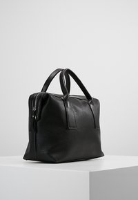 Svart texturerad läderhandväska med två handtag och en silverfärgad dragkedja, vilande på en vit pelare mot en plain grå bakgrund.