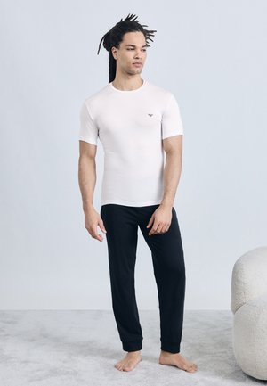 Hombre con rastas descalzo, vistiendo una camiseta blanca ajustada y pantalones negros, frente a un fondo gris claro liso.