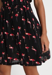 Jupe noire avec petits motifs de flamants rouges, taille froncée, et tissu léger texturé. Elle présente un design fluide et une coupe mi-longue.