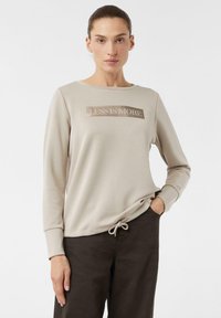 Jasno beżowy sweatshirt z długim rękawem, z błyszczącym napisem "LESS IS MORE" z przodu oraz detalem w postaci sznureczka u dołu.