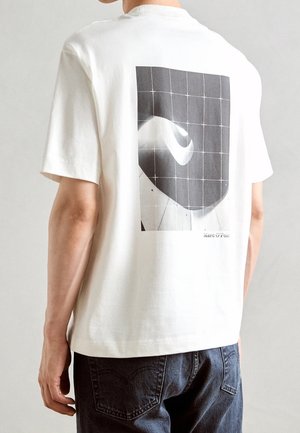 Osoba ubrana w biały t-shirt z czarno-białym abstrakcyjnym wzorem kratki i spirali na plecach oraz ciemne jeansy.