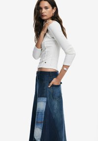 Weißes, geripptes Langarmoberteil kombiniert mit einem Patchwork-Denim-Maxirock in verschiedenen Blautönen und mit Taschen. Insgesamt ein minimalistisches Design.