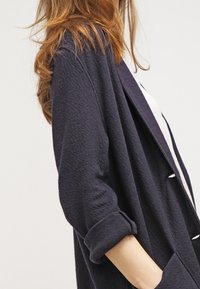 Námorně modrý texturovaný cardigan s uvolněným střihem, ohrnutými rukávy a otevřeným předním designem. Látka vypadá měkce a neformálně, ideální pro vrstvení.