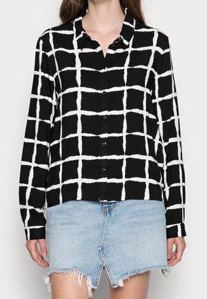 Femme portant une chemise noire à boutons avec un motif à carreaux blancs et une mini-jupe en denim effilochée de couleur bleu clair.