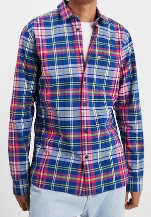 Mand iført en blå, pink og gul ternet skjorte med knapper over en hvid t-shirt og lyseblå jeans, stående foran en hvid baggrund.