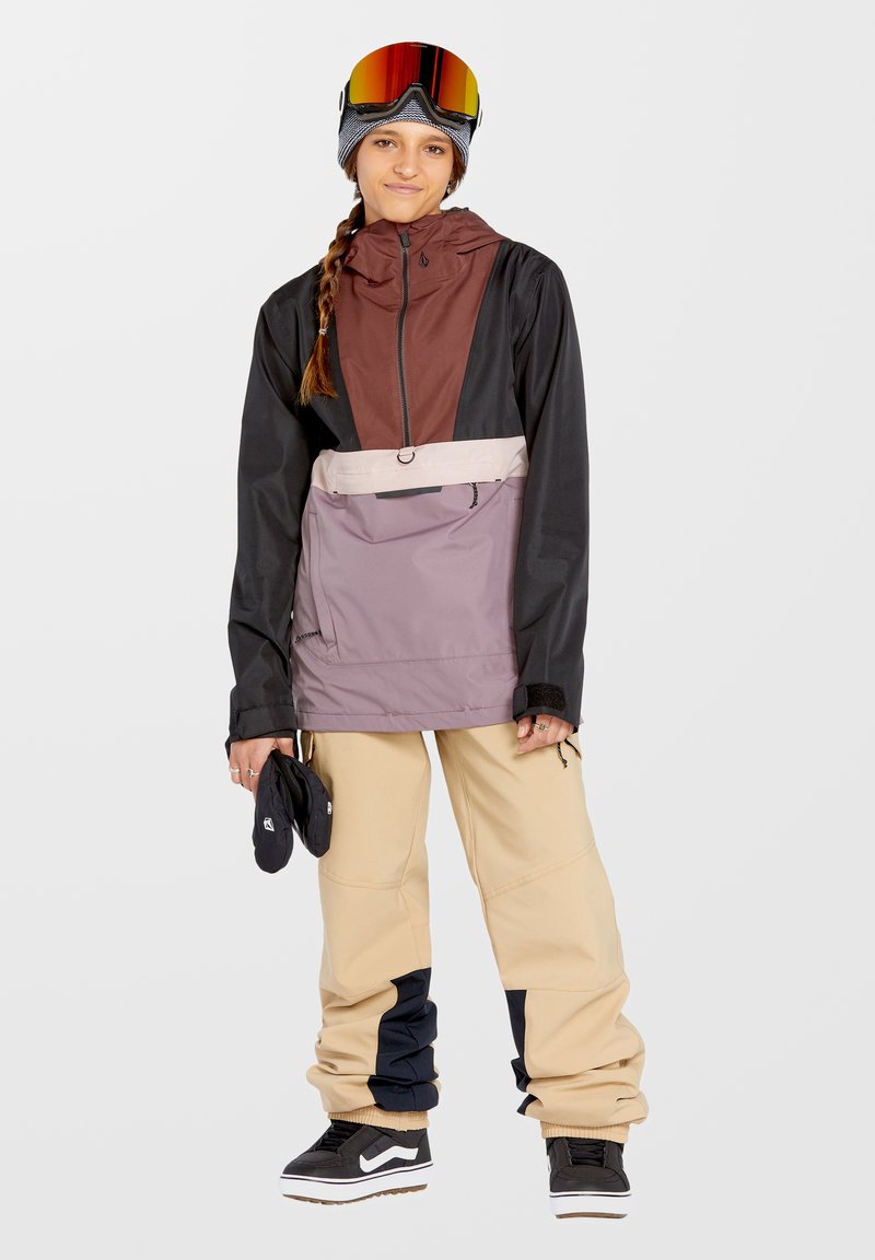 Schneeboardjacke in Schwarz, Burgunderrot und Violett mit einem halben Reißverschluss; kombiniert mit beigen Hosen, die schwarze und marineblaue Akzente haben, und schwarzen Schuhen.