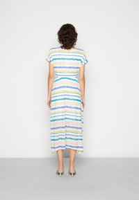 Femme aux cheveux bouclés portant une robe blanche mi-longue à rayures horizontales multicolores, debout devant un mur blanc uni.