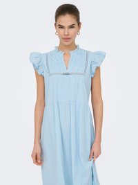 Femme portant une robe sans manches bleu clair avec des épaules volantées et un décolleté carré plissé, debout devant un fond uni.