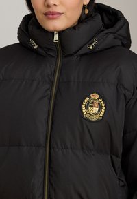 Juoda pūkinė striukė su užtrauktuku ir gobtuvu. Kairėje krūtinės pusėje yra auksinis emblemų su karūna ir laurų detale.