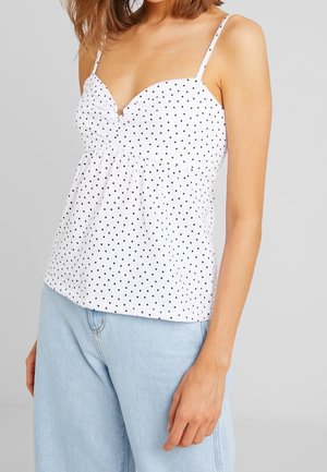 Mujer con top blanco de tirantes finos con lunares negros y vaqueros de tiro alto azul claro frente a un fondo gris.