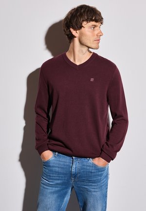 Burgunderfarbener V-Ausschnitt-Pullover mit langen Ärmeln, aus einem glatten Material gefertigt. Kombiniert mit hellblauen Jeans, die ein einfaches, unaufgeregtes Design zeigen.
