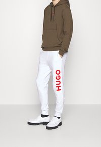 Brun hættejakke med en frontlomme og snøre. Hvide sweatpants med en markant rød "HUGO" tryk på siden. Hvide sko med sorte såler.