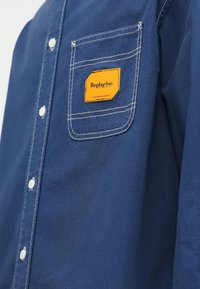 Μπλε πουκάμισο με κουμπιά από βαμβακερό μείγμα, διαθέτει τσέπη με πορτοκαλί ετικέτα και λευκή ραφή. Κλασικό γιακά και στρογγυλεμένο τελείωμα.