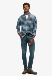 Camicia di flanella a quadri blu con colletto, T-shirt bianca sotto, abbinata a jeans blu e stivali marroni con lacci. Design semplice e casual.