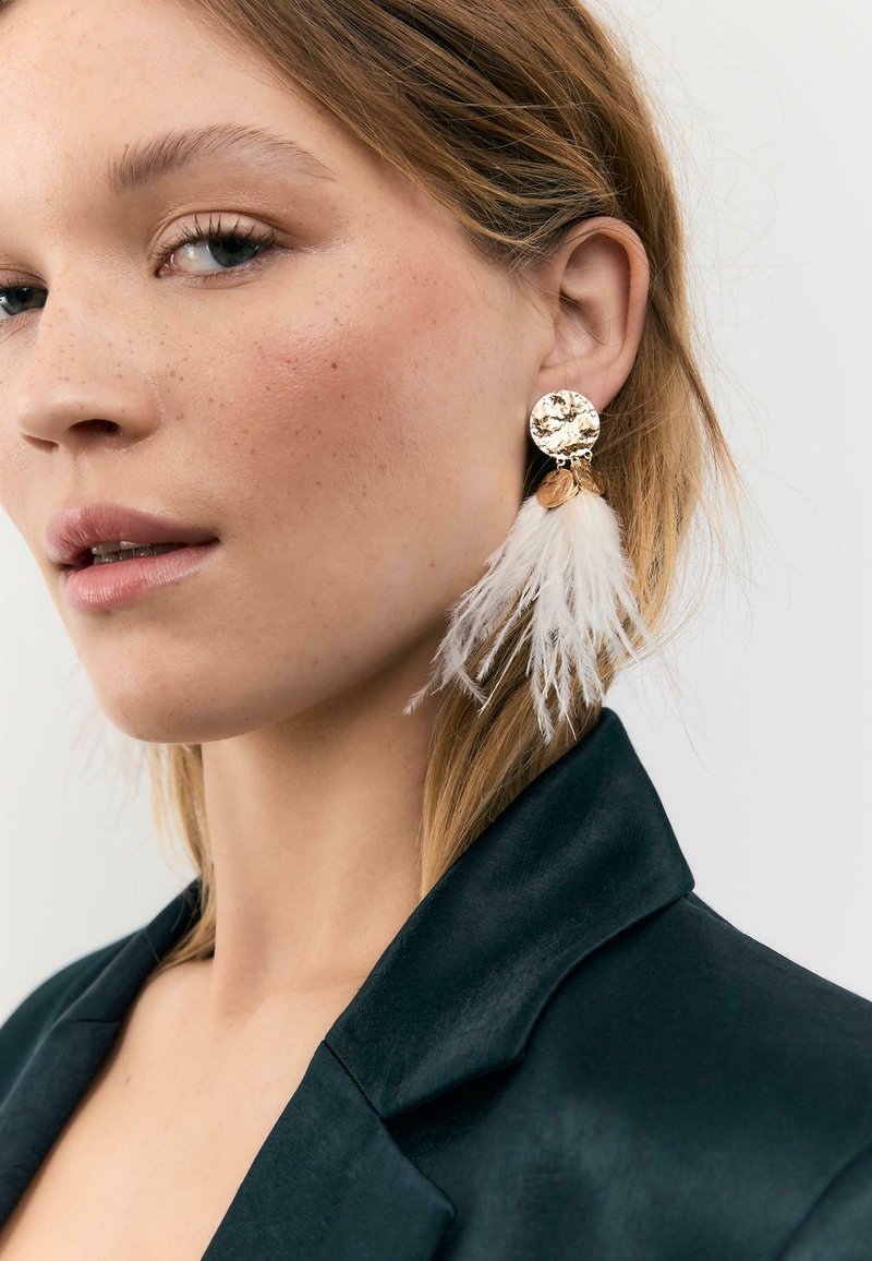 Jeune femme à la peau claire et parsemée de taches de rousseur, portant une grande boucle d'oreille en or ornée de plumes blanches et un blazer noir, sur un fond uni.