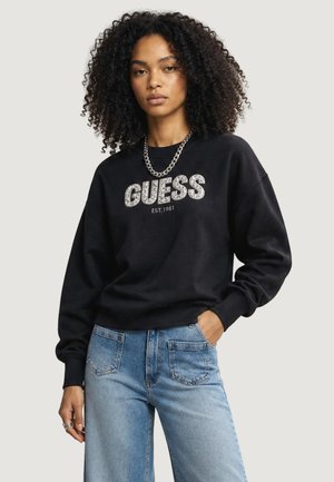 Kvinde med krøllet hår iført en sort Guess sweatshirt med hvid tekst og lyseblå jeans, stående med den ene hånd i lommen.