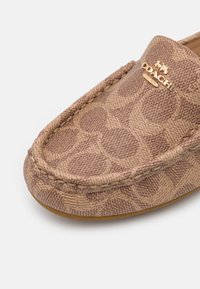 Chaussure slip-on marron à motif avec un tissu texturé, présentant un bord cousu et un accent logo doré sur le dessus. La forme est plate et arrondie.