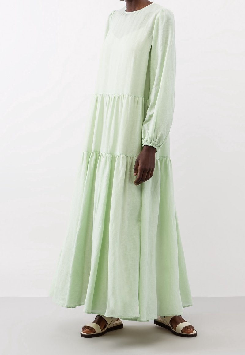 Femme portant une longue robe à volants vert clair avec manches longues et des sandales blanches plates, debout devant un fond blanc uni.