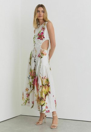 Žena oblečená v bielych maxi šatách bez rukávov s kvetinovým vzorom a bočnými výstrihmi, doplnená o béžové sandále na vysokom opätku.