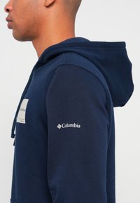 Profil osoby v námorní modrej mikine Columbia s bielym logom na rukáve a na hrudi na jednoduchom pozadí.