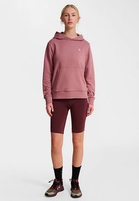 Lichtroze hoodie met een voorzak en logo, gecombineerd met bordeauxrode fietsbroek en donker schoeisel met rode accenten.