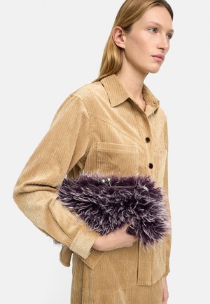 Kobieta w beżowej koszuli ze sztruksu trzymająca fioletową puszystą kopertówkę, zwrócona w prawo na tle jednolitego jasnego tła.