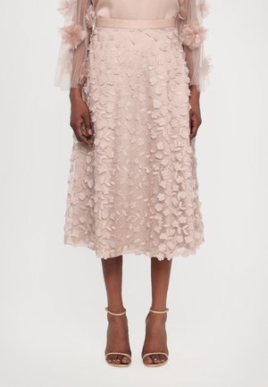 Kvinde iført en knælængde blush pink nederdel med blomsteret blonderbroderi og gennemsigtig blomstret ærmet top, kombineret med nøgne ankelrem hæle.