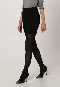 Falda negra texturizada combinada con medias opacas negras y tacones de punta con acentos de púas. Una blusa gris de manga larga completa el conjunto.